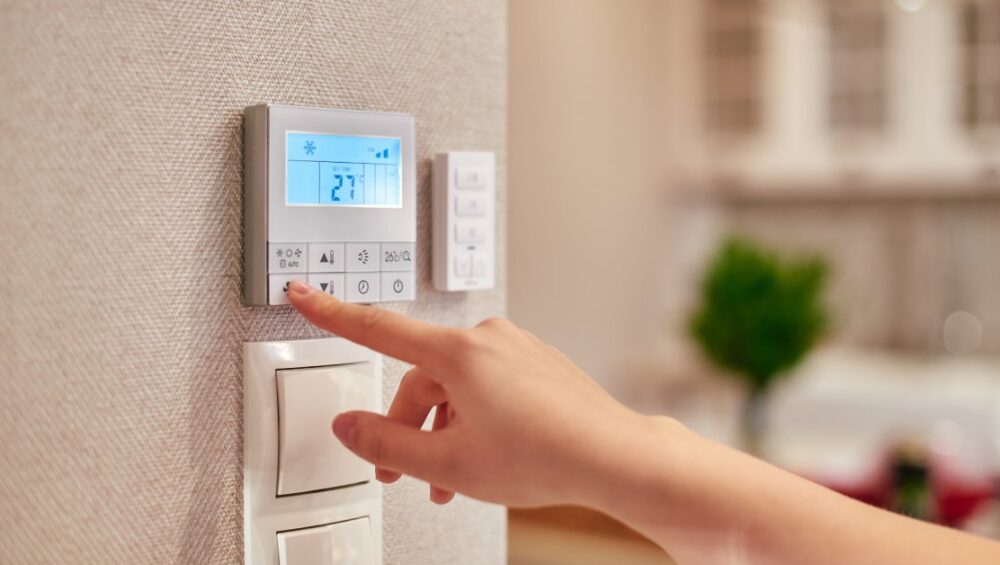 A person’s hand reaching towards a thermostat on a wall in the kitchen of a home to change the fan speed.