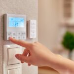 A person’s hand reaching towards a thermostat on a wall in the kitchen of a home to change the fan speed.