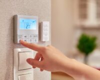 A person’s hand reaching towards a thermostat on a wall in the kitchen of a home to change the fan speed.