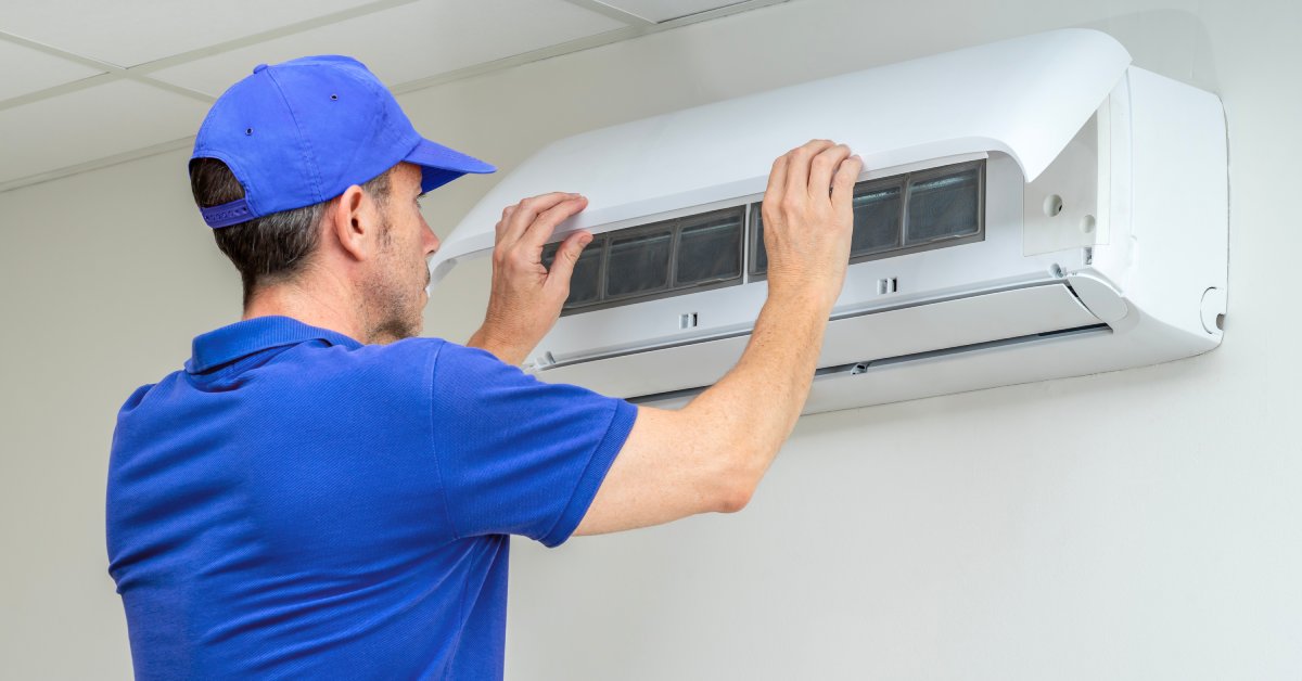 A repair technician wearing a blue shirt and hat putting the cover back on an air conditioning unit on a wall.