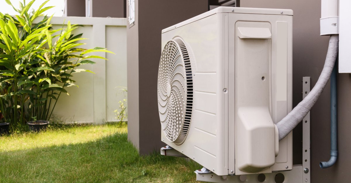 An air conditioning unit mounted on the outside wall of a home. There’s a fence with a plant in front of it beneath a blue sky.