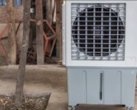 An evaporative cooler on a cart with wheels. The cooler is located on a sidewalk next to a small tree.