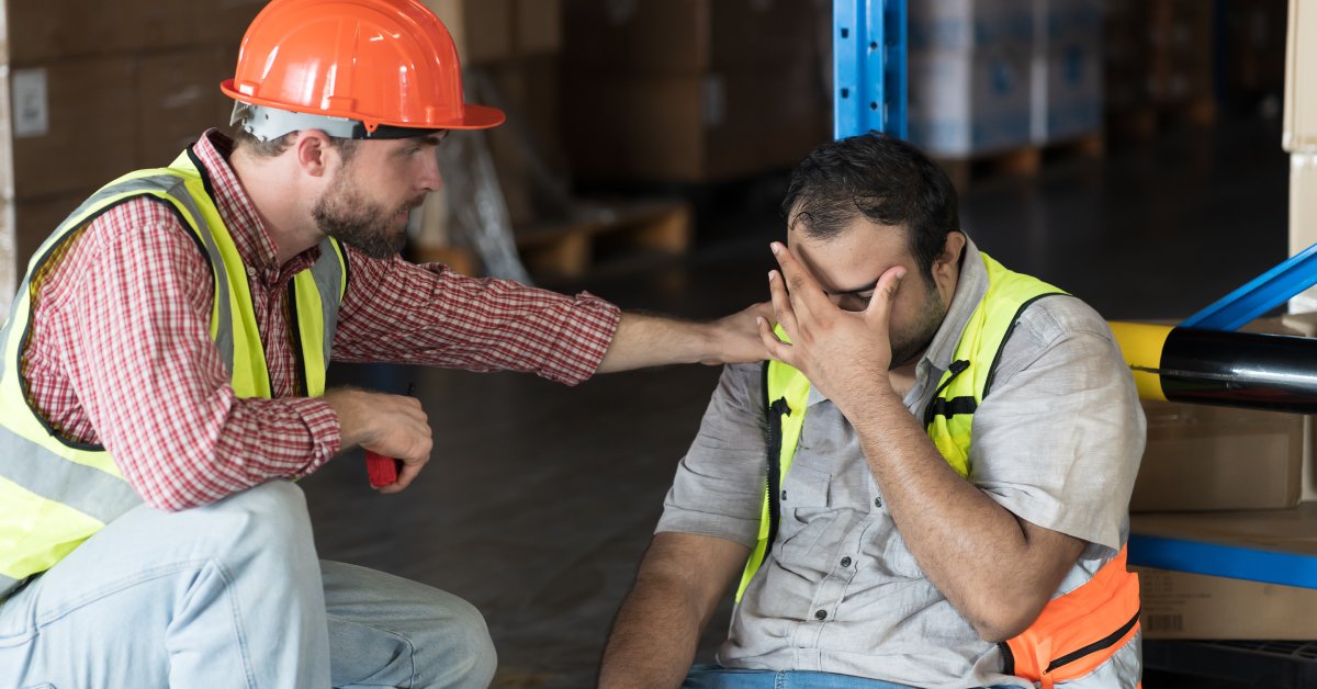 A man sitting down on a factory floor with his hand over his face. Another man his kneeling down with a hand on his shoulder.