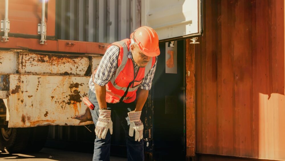 A man wearing a high-visibility vest and a hard hat holding his hands on his knees in front of a shipping truck.