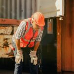 A man wearing a high-visibility vest and a hard hat holding his hands on his knees in front of a shipping truck.