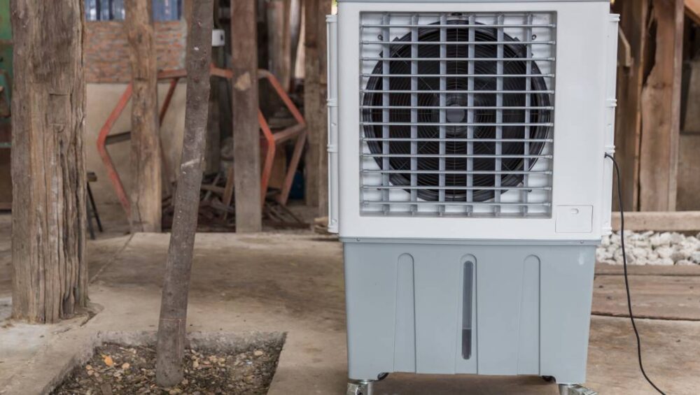 A portable evaporative cooler on wheels sitting outside on the concrete next to a small tree and a row of wooden posts.