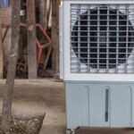 A portable evaporative cooler on wheels sitting outside on the concrete next to a small tree and a row of wooden posts.