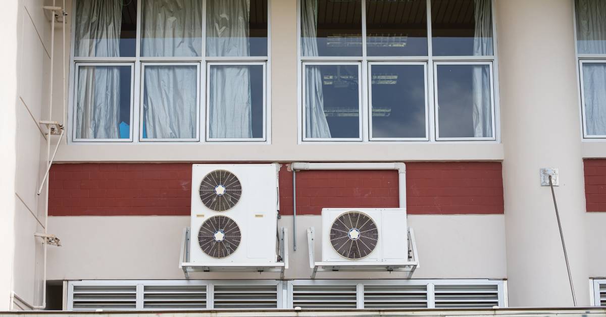 A building with a red stripe and windows with curtains. There’s a set of cooling fans attached to the side of it.