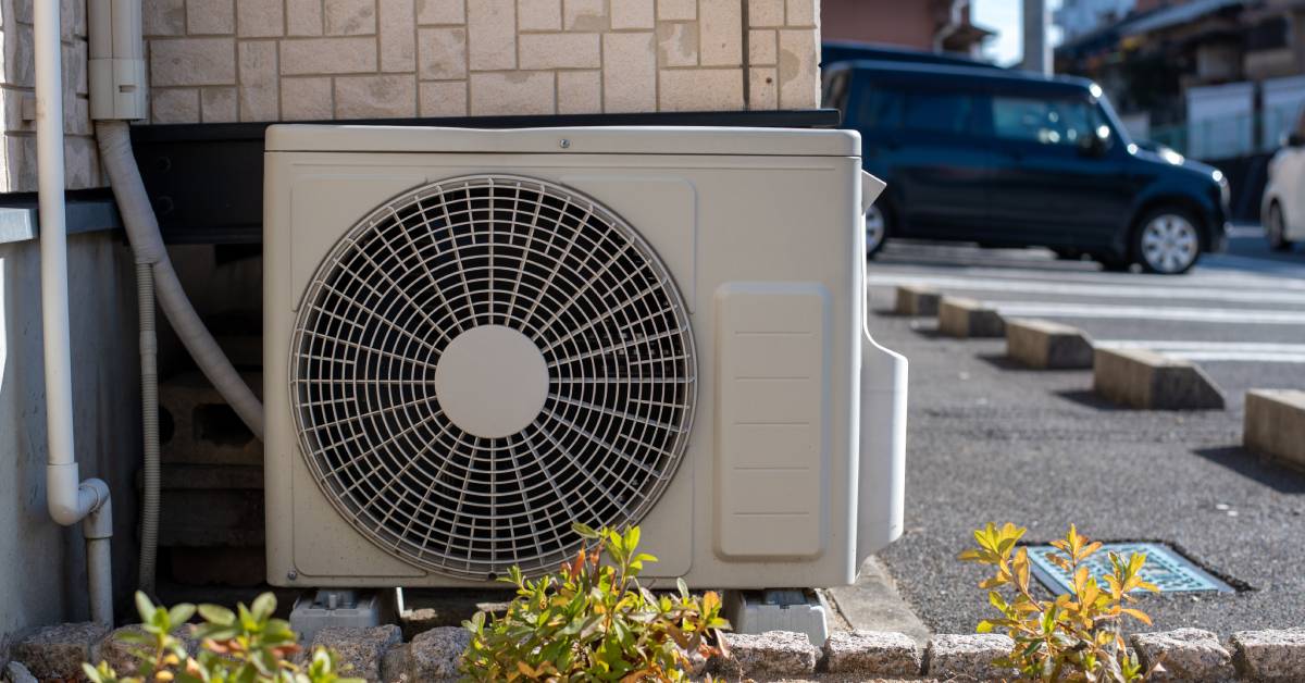 A cooling unit positioned outside of a building next to a small garden and a parking lot with vehicles in it.