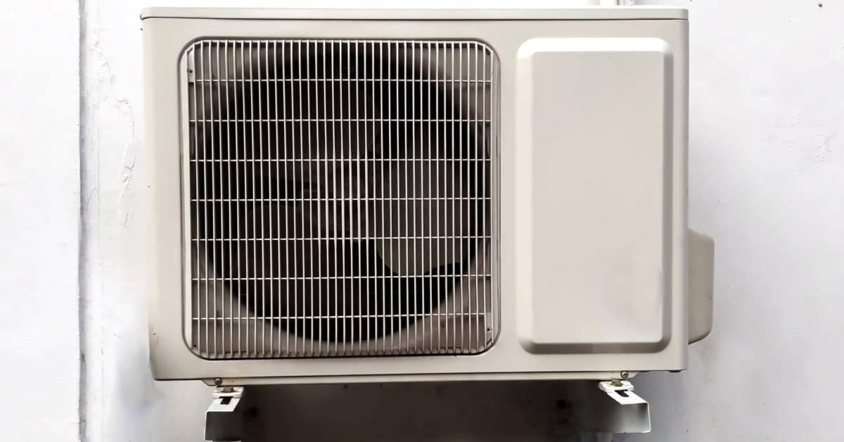 A tan-colored swamp cooler installed on a mount on the side of a cement building. The cooler has wires attached.