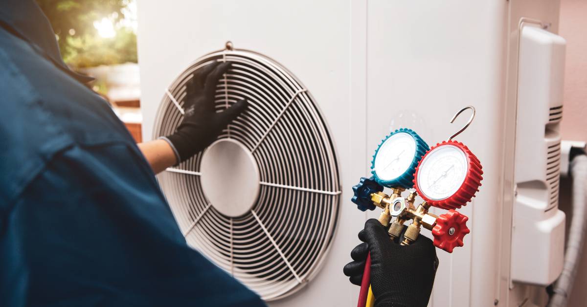 A worker wearing a blue uniform and gloves holds a component while performing maintenance on an outdoor HVAC system.