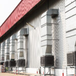 A long row of evaporative cooling units on top of metal stands connected to airducts outside of a building.