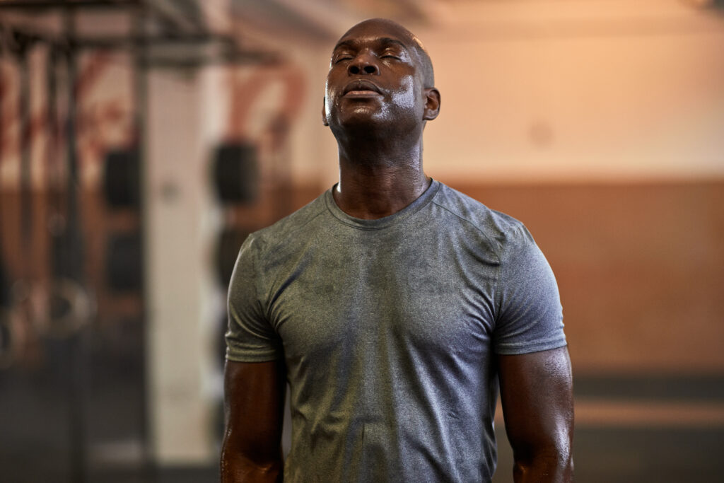 A man wearing a sweat-covered gray t-shirt standing in a gym looking up with his eyes closed in front of exercise equipment.