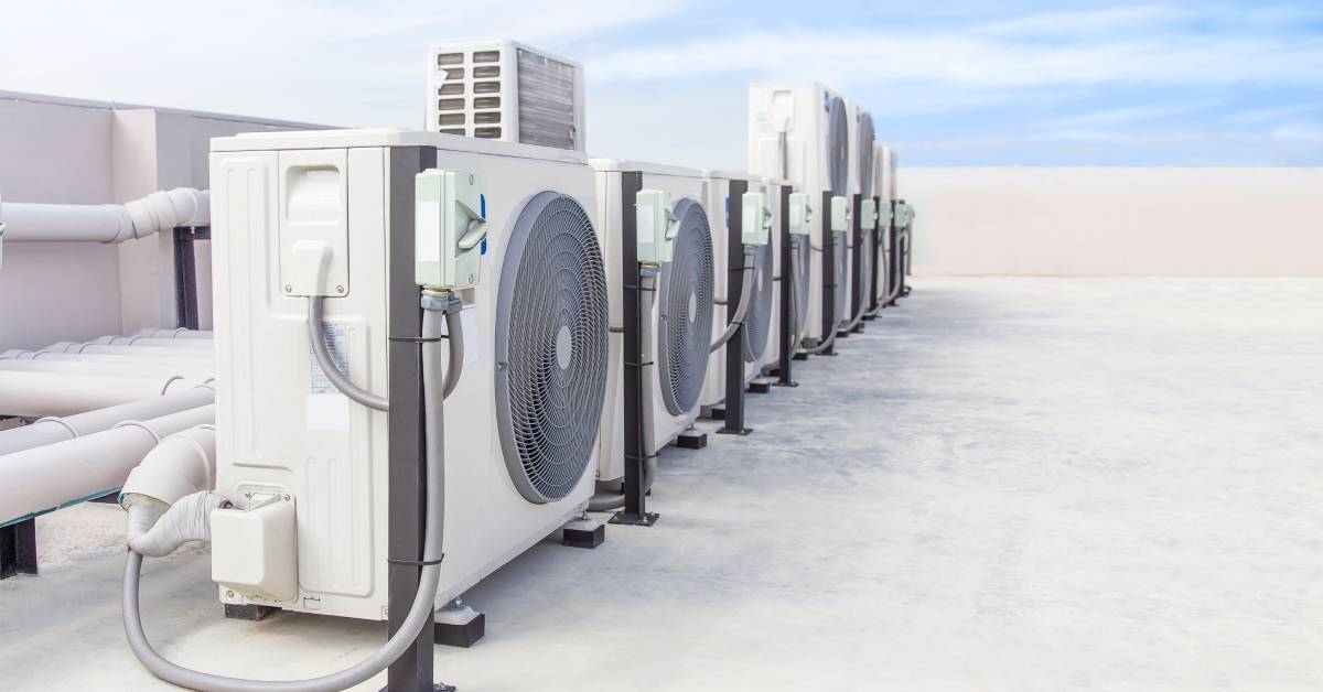 A row of white evaporative coolers on the roof of an industrial building. Blue sky and clouds are visible behind the coolers.